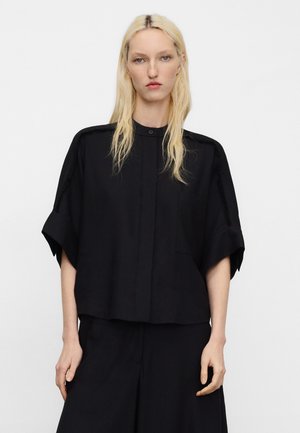 Blonde vrouw met steil haar draagt een loszittend zwart T-shirt met korte mouwen, verborgen knopen en bijpassende zwarte broek.