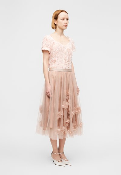 Ανοιχτό ροζ floral τοπ με κοντά μανίκια, σε συνδυασμό με διάφανο, στρωματοποιημένο τούλινο φούστα διακοσμημένη με τριανταφυλλένια εφαρμογή, και λευκά παπούτσια με χοντρό τακούνι.