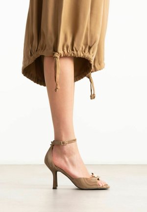Damesbeen met een beige sandalen met hak en enkelbandje, gecombineerd met een lichtbruine rok met verstelbare koordjes aan de zoom.
