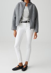 Femme portant une veste grise zippée, un haut gris clair, un pantalon blanc ajusté avec une ceinture noire, et des chaussures plates noires à bride boucle, debout devant un fond uni.