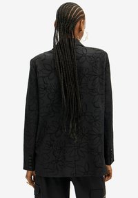 Osoba s dlouhými zapletenými vlasy, která nosí velké kruhové náušnice a černý blazer s jemným květinovým vzorem, viděná zezadu.
