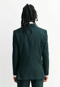 Donkergroen blazer van gestructureerde stof. Kenmerkt zich door een getailleerde pasvorm, met een reverskraag en knoopmanchetten, en heeft een gladde achterkant.
