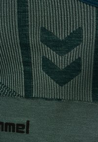 Zielona, prążkowana tkanina w wzór zygzakowy w ciemniejszych odcieniach. Posiada teksturowane wykończenie oraz czarne logo marki na dole.