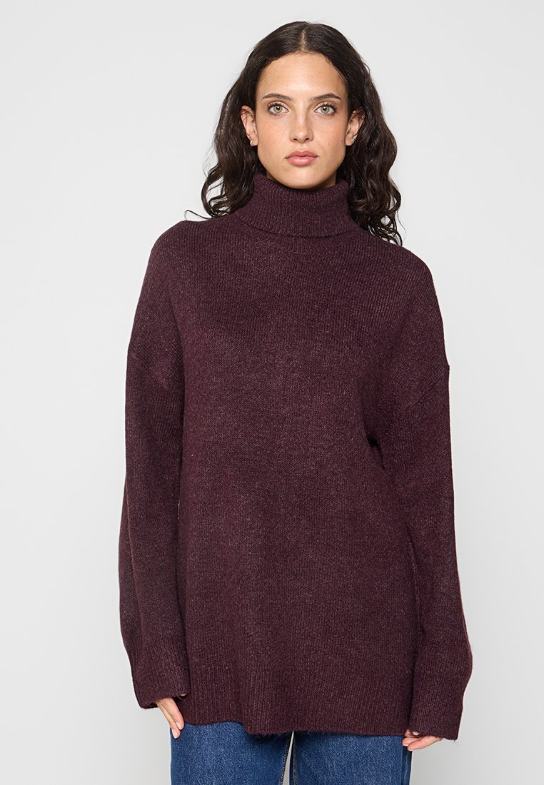 Pull col roulé bordeaux à coupe décontractée, texture côtelée et épaules tombantes, associé à un jean en denim bleu.