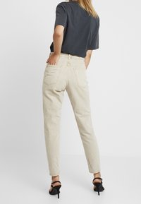 Pantalones beige de talle alto con un corte relajado, que cuentan con bolsillos traseros y un acabado texturizado, combinados con una camiseta oscura y tacones negros.