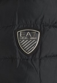 Černý prošívaný kabát z tkaniny s výraznou kovovou nášivkou loga EA7 ve tvaru štítu a detailem sepraného proužkovaného vzoru.