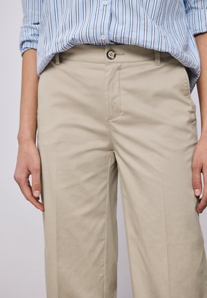 Osoba ubrana w jasnobeżowe spodnie na guziki i niebiesko-białą koszulę w paski wpuszczoną do środka, z rękami swobodnie opuszczonymi wzdłuż ciała.