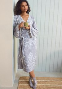 Accappatoio in raso grigio con motivo floreale, dettagli in pizzo e cintura. Modella che tiene una tazza, indossando calzini floreali coordinati, in un ambiente interno morbido.