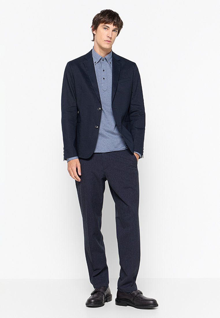 Hombre vestido con un blazer azul marino, camisa de polo azul de botones, pantalones oscuros y zapatos negros, de pie con una mano en el bolsillo, contra un fondo blanco.