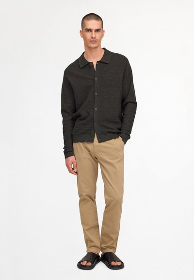 Homme debout portant un cardigan noir boutonné, un pantalon beige et des sandales noires à enfiler sur fond blanc.
