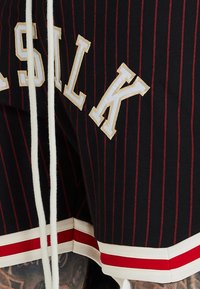 Svarta sportshorts med röda ränder och vit text. Har en vit dragsko och detaljer med röd-vit rand vid fållen.