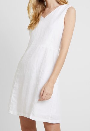Mujer vistiendo un vestido blanco sin mangas con escote en V y detalles sutiles de costuras, de pie frente a un fondo claro y liso.