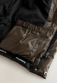 Mörkbrun puffjacka med svart innerfoder. Utrustad med en zippered ficka, elastisk midja och en märkt svart etikett vid fållen.