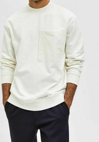 Vitt sweatshirt gjort av bomull med rund halsringning, ribbade ärmslut och en texturerad framficka. Kombinerat med mörkfärgade byxor.