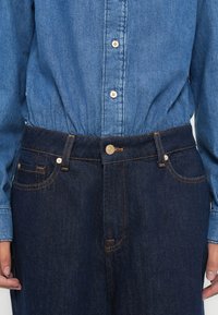 Τζιν πουκάμισο σε ανοιχτό μπλε με κουμπιά μπροστά και στήσιμο στη μέση· συνδυάζεται με σκουρόχρωμο μπλε τζιν παντελόνι με πορτοκαλί ραφές και τσέπες.