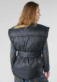 Gilet noir matelassé avec des épaules larges et une taille ceinturée, affichant une texture lisse et brillante ainsi qu'un design matelassé au dos.