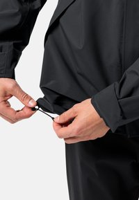 Veste noire résistante aux intempéries avec ourlet réglable à cordon. Présente une finition lisse et mate ainsi que des poignets ajustés. Des mains montrent l'ajustement.