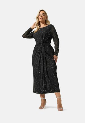 Mujer con un vestido largo negro con pequeños lunares blancos, mangas largas y sandalias plateadas de tacón alto, de pie con una mano tocándose el cabello.