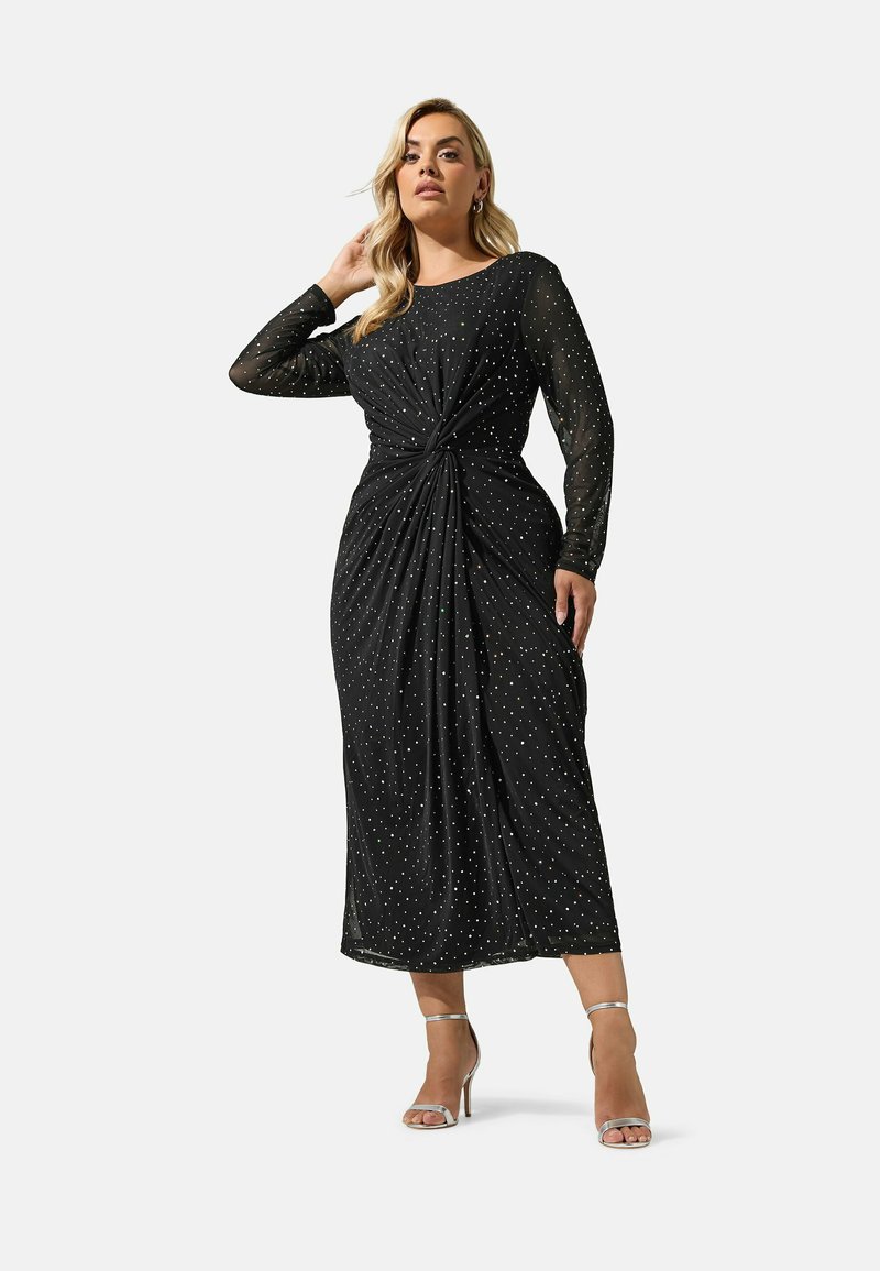 Femme portant une longue robe noire à petits pois blancs, manches longues, et des sandales argentées à talons hauts, debout, une main touchant ses cheveux.