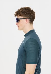 Sportsskjorte i mørk teal med teksturerede ærmer og kort krave. Modellen bærer oversized lilla solbriller. Glat stof, figursyet design.