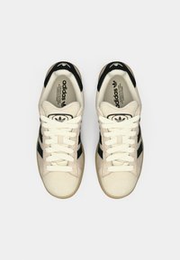 Beigesmockade sneakers med svarta ränder, rund svart logotyp på tungan och krämfärgade skosnören. Flat gummisula och texturerad ovandel.