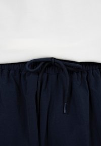 Balti marškiniai įkišti į tamsiai mėlynas kelnes su elastinga juosmens juosta ir surištu raišteliu centre.