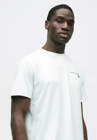 Biała koszulka z krótkim rękawem, okrągłym dekoltem i subtelnym zielonym logo Lacoste na lewej piersi. Miękki, gładki materiał.