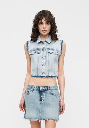 Giovane donna che indossa una giacca di jeans senza maniche corta e una gonna di jeans con bottoni sul davanti e bordi sfrangiati, in piedi davanti a uno sfondo semplice.