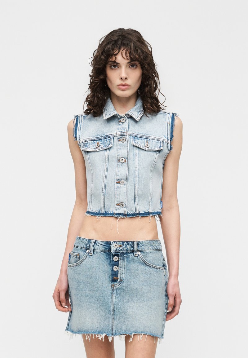 Νεαρή γυναίκα που φοράει ένα αμάνικο κοντό τζιν μπουφάν και μια τζιν φούστα με κουμπιά μπροστά και ξεχωριστές άκρες, στέκεται μπροστά σε ένα απλό φόντο.