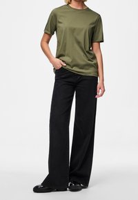 Olivově zelené bavlněné tričko s ohrnutými rukávy, v kombinaci s širokými černými džínami. Modelova ruka v kapse, boty s lesklým povrchem.