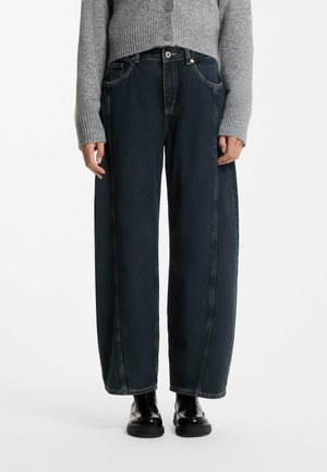 Persoon draagt donkere wijde jeans, een grijze trui met knopen en zwarte glanzende schoenen en staat tegen een witte achtergrond.
