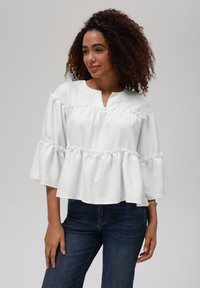 Hvit peplum-top med volangdetaljer, med halv knapplås og brede ermer. Båret med mørkeblå jeans, som viser et strukturert stoff.