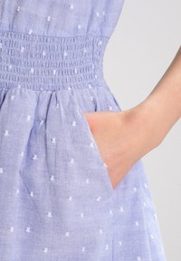 Vaaleansininen mekko, jossa on valkoisia pilkkuja, varustettu smockattulla vyötäröllä ja sivutaskulla. Kangas on kohokuvioitu ja kevyt.