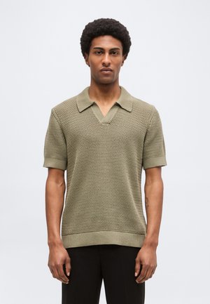 Młody mężczyzna z krótkimi kręconymi włosami, ubrany w luźną oliwkowozieloną dzianą koszulkę polo z krótkim rękawem i czarne spodnie, stojący na jednolitym tle.