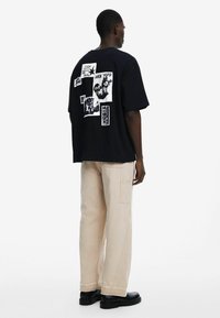 Black oversized t-shirt featuring graphic patches in white, with short sleeves and a round neckline. Paired with beige cargo pants and black boots.
