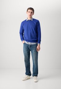 Felpa blu con polsini a coste, indossata sopra una camicia a righe con colletto, abbinata a jeans blu e sneaker bianche. Strato semplice e casual.