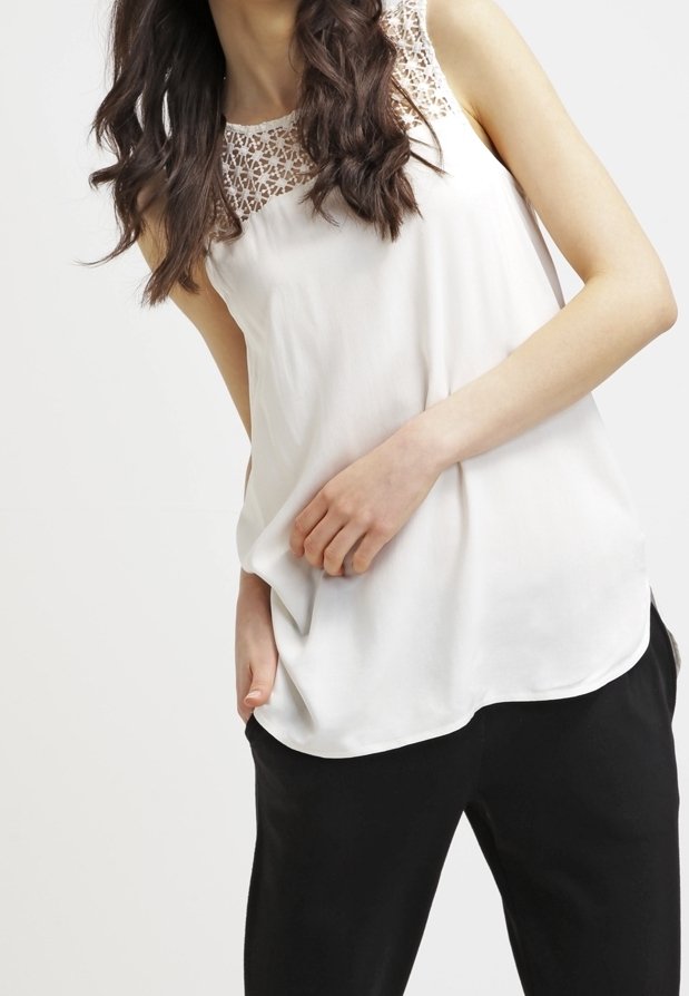 Femme portant un haut blanc sans manches avec un détail en dentelle et un pantalon noir, posant avec une main dans la poche et l'autre tenant le haut.
