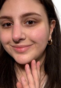 Lähikuva keskivärisestä ihosta, tummista hiuksista ja kultaketjuisista korviksista. Huulilla on kevyttä sävyä ja kädet lepäävät leualla.