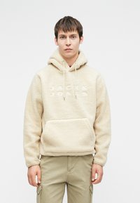 Νέος άντρας που φοράει μια κρέμα φλις ζακέτα με μπροστινή τσέπη και κουκούλα με κορδόνι, συνδυασμένη με ανοιχτό καφέ cargo παντελόνι σε απλό φόντο.