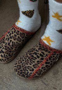 Træsko med leopardprint og rød syning, båret med hvide, bløde sokker med gule stjerner og sorte trekanter, på en tekstureret overflade.