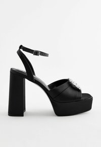 Black satin platform sandals with a chunky heel, featuring a wide strap and a decorative jewel embellishment on the front.