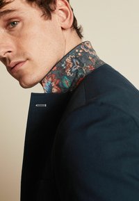 Mand i navy blazer med farverigt blomstermønstrede krave oppe, der ser let over sin skulder imod en ensfarvet baggrund.