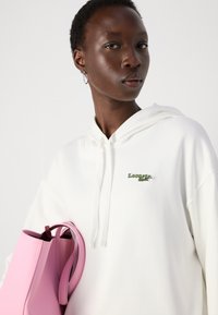 Vit hoodie med en grön broderad logotyp, i kombination med en strukturerad rosa väska som visar en slät textur och kontrasterande färger.