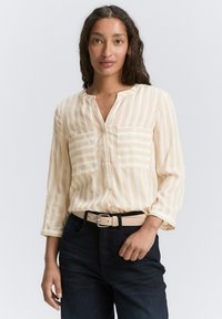 Lys gul stribet skjorte med knapper, to forlommer, lange ærmer og blødt stof, parret med mørke jeans og et beige bælte.
