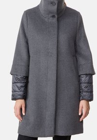 Šedý kabát s kostkovaným vzorem houndstooth, vysokým límcem, dvěma knoflíky a prošívanými stříbrnošedými rukávy. Materiál se zdá být měkký a strukturovaný.