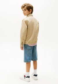 Veste légère beige, shorts en denim et baskets blanches. La chemise en dessous a des rayures rouges et blanches. Cheveux courts, vu de dos.