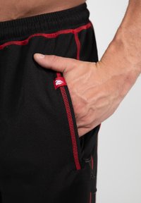 Zwarte shorts met rode stiksels, elastische tailleband en zijzak. Materiaal lijkt gestructureerd, wat de ademendheid en het comfort verbetert.