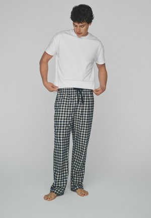 Wit T-shirt met korte mouwen gecombineerd met marineblauwe en crèmekleurige geruite pyjamabroek met een elastische tailleband en trekkoord. Blootsvoets.