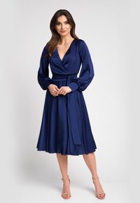 Granatowa sukienka kopertowa z satynowego materiału, z długimi bufkami, głębokim dekoltem w serek oraz przewiązywanym paskiem w talii. Rozkloszowana spódnica sięgająca do połowy łydki.