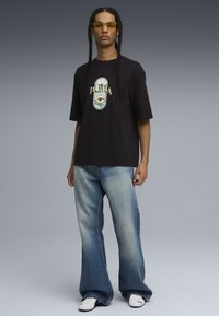 Sort bomulds T-shirt med et farverigt grafisk logo; sammen med lyseblå flared jeans; modellen har gule solbriller og hvide sko på.
